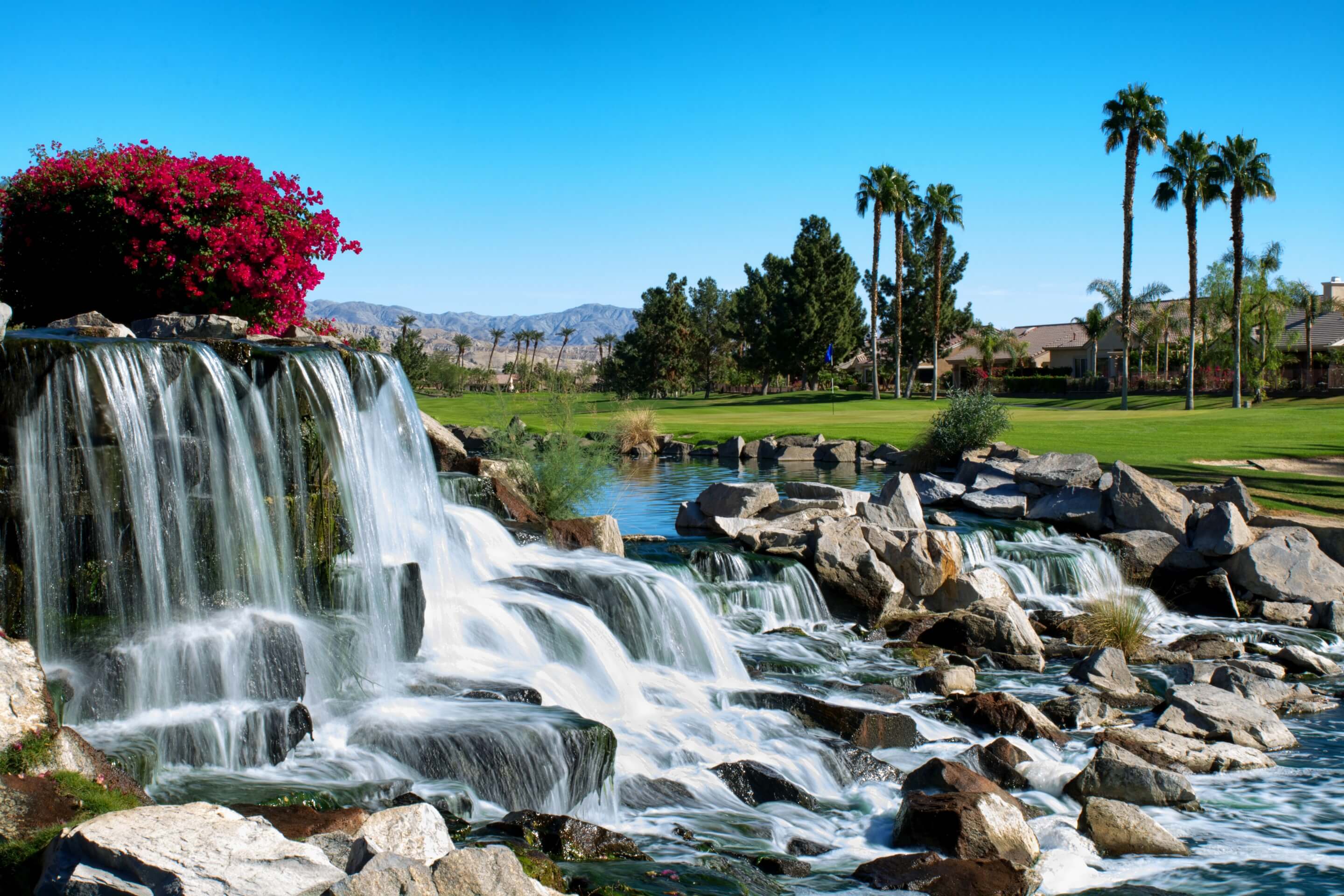 Waterfalls, palms and landscaped grounds at Sun City Palm Desert, with mountains in the distance.